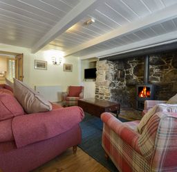 A living room with sofas and armchairs around a wood burning stove set in a stone fireplace at April Cottage in Coverack