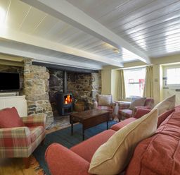 A living room with a stone fireplace wood stove red and plaid armchairs a red sofa a wooden coffee table a television on a stand and a door with windows at April Cottage in Coverack