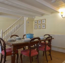 A dining room with a wooden table and six red cushioned chairs near a staircase at April Cottage in Coverack