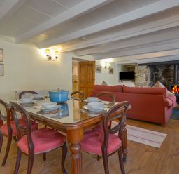 A dining area with a wooden table set with bowls and cutlery and a blue pot next to a living room with a red sofa and a fireplace at April Cottage in Coverack