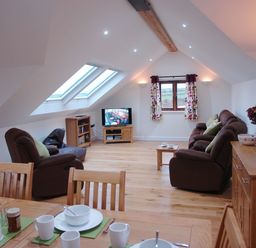 An attic living room with sofas a television dining table set and flowers on a cabinet at The Hayloft in Breage