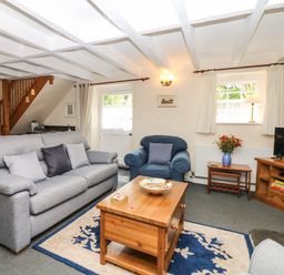 A living room with gray sofa, blue armchair, wooden coffee table, rug, TV on wooden stand, flowers on a side table at Badger Cottage in Mawgan-in-Meneage near Helston