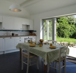 A kitchen with white cabinets and a dining table set with dishes next to a large open door showing a garden at The Studio in Rinsey Cove near Praa Sands