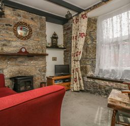 A sitting room with stone walls a fireplace a red sofa a wooden chair and a window with floral curtains at Mollys Cottage in St Just
