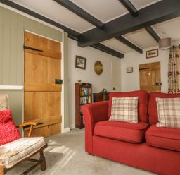 A living room with a red sofa and plaid cushions next to wooden doors and a wooden chair with a red cushion at Mollys Cottage in St Just