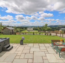 A patio with a hot tub and a table with benches overlooking a grassy yard and hills at Hillside View in Rhayader