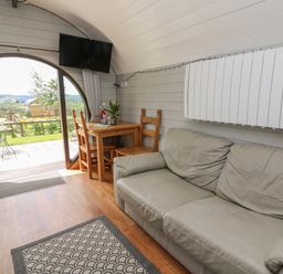 An interior room with a leather sofa wooden table with chairs flat screen tv and circular glass door showing outdoor seating at Woodland View in Rhayader