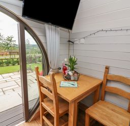 A small wooden dining table with two chairs near a round glass door and a mounted TV at Woodland View in Rhayader