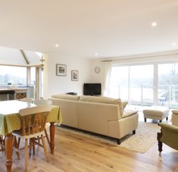 An open living room and dining area with a dining table covered with a green polka dot tablecloth chairs beige sofas an armchair a small TV and large windows with curtains at Treetops in Golant near Fowey