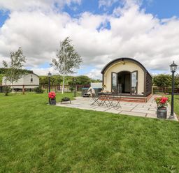 A small cabin with an open door on a stone patio with table and chairs and flowers on grass at Mountain View in Rhayader
