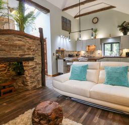 A living room with a cream sofa and turquoise cushions next to a wooden coffee table and a kitchen with grey cabinets at Elvan Cottage in Liskeard