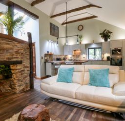 An open living room and kitchen area with a white sofa and blue cushions a wooden coffee table exposed wooden beams and stone wall at Elvan Cottage in Liskeard
