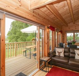 A wooden living room with a brown sofa and deer pillows next to a glass door leading to a deck with a picnic table at Sunnyside Lodge in Crewkerne