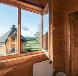 A wooden interior with an open window showing a wooden cabin and green fields outside at Sunnyside Lodge Crewkerne