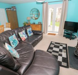 A living room with a brown leather sectional sofa and patterned cushions a wooden dining table with chairs a wooden cabinet a round mirror and a television on a stand at 4 Hanbury Lodge Pentlepoir
