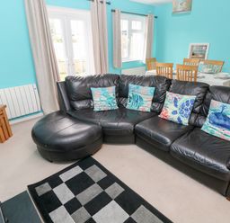 A living room with a black leather sectional sofa with sea-themed pillows a dining table with wooden chairs and tableware blue walls and beige curtains at 4 Hanbury Lodge in Pentlepoir