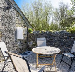An outdoor patio with a round wooden table and four mesh chairs by a stone wall and house at Moor View Chapel in Camelford