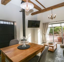 A dining area with a wooden table, chairs, a wall-mounted television, and a sitting area with a coffee table and sofa with sliding glass doors to a patio at The Stables in Cross Keys near Hereford