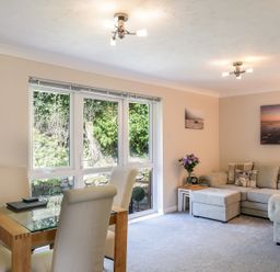 A living room with a glass dining table and white chairs next to a beige sofa with cushions and a side table with flowers at Little Retreat in Liskeard