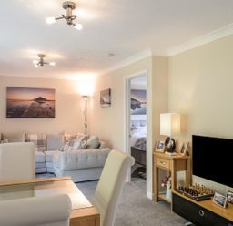 A living room with a grey sectional sofa a glass dining table with chairs a wooden TV stand with a television and framed photos at Little Retreat in Liskeard