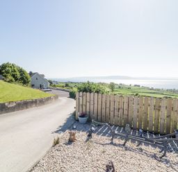 A rural driveway with a gray house and a gravel garden area with decorative figurines near a wooden fence at Tullyally in Redcastle