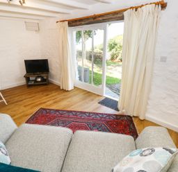 A living room with a gray sofa and patterned cushions a red and gray rug a small TV on a wooden stand and sliding glass doors with cream curtains at The Granary Cottage in Llangrannog near Cardigan