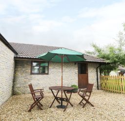 An outdoor patio area with a round wooden table and three wooden chairs under a green umbrella next to a stone wall building and wooden fence at The Old Goat Barn at Trout Cottage in Somerton