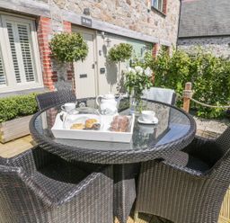 An outdoor patio area with a round glass table and four wicker chairs a tray with teapot scones and cups and a vase with flowers in front of a stone building at Oh Buoy Charlestown