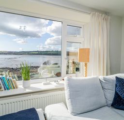 A room with a large window overlooking a body of water with a sofa lamp and bookshelf by the window at Clamoak Cottage in Clamoak near Bere Alston