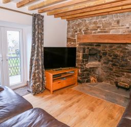 A living room with a stone fireplace two leather sofas a wooden TV stand with a television and glass doors with curtains at Crooked Hill Cottage in Ammanford