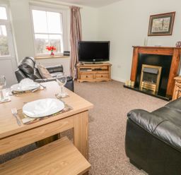 A living room with a wooden dining table set for two black leather sofas a wooden fireplace and a television at 2 Park Nook Close in Waberthwaite
