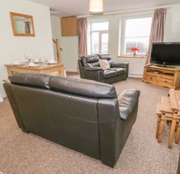 A living room with black leather sofas a wooden TV stand with a television a dining table set and a side table with a lamp at 2 Park Nook Close in Waberthwaite