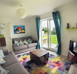 A living room with two sofas and colorful cushions a wooden coffee table a multicolored rug a TV with speakers and a sliding glass door with blue curtains at Driftwood in Bude