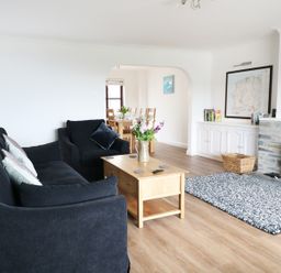 A living room with a black sofa and armchair a wooden coffee table and a fireplace a dining area visible through an archway at Seal Cottage in Mortehoe