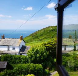 View of a white house with chimneys surrounded by green vegetation and hills with the sea in the background at Seal Cottage in Mortehoe