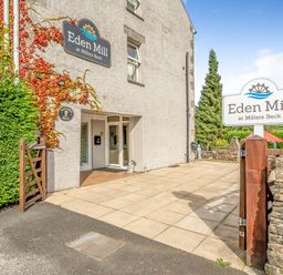 The entrance of Eden Mill at Millers Beck with signs, a wooden gate, a tiled patio, and surrounding greenery in Kendal