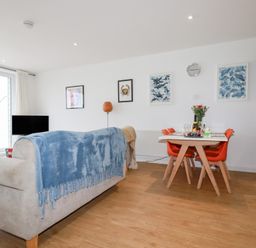 A living area with a beige sofa covered by a blue throw facing a television and a dining table with four orange chairs at Buena Vista in Newquay