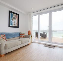 A living room with a beige sofa, colorful cushions, a framed wall art, large glass sliding doors leading to an outdoor seating area with a view of the sea at Buena Vista in Newquay