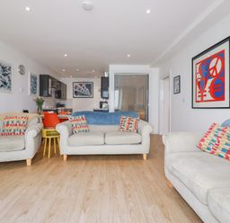 A living room with two sofas and an armchair with patterned cushions and blue throws and a kitchen area with black cabinets and a dining table with orange chairs at Buena Vista in Newquay