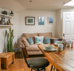 A small living area with a sofa pillows dining table with dishes and mugs wooden stools and shelves with decorations at The Compass in Swanage