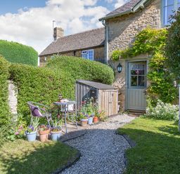A garden with a gravel path leading to a door of a stone house with potted plants and a small table with chairs at Puffitts Cottage in Bourton-on-the-Water