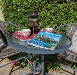 A glass outdoor table with a French press coffee maker two cups and a magazine between two wicker chairs with floral cushions at Puffitts Cottage Bourton-on-the-Water