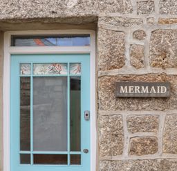A stone exterior wall with a blue door and a sign that reads Mermaid at Mermaid Cottage in Mousehole