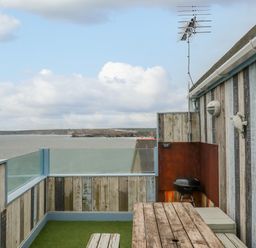 A rooftop patio with wooden walls glass railing a wooden table benches a barbecue grill and a view of the sea at Sailor's Rest in Newquay