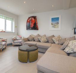 A living room with a beige sectional sofa and two armchairs with text patterns a round table and wall art at Sailor's Rest in Newquay