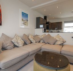 A living area with a sectional sofa and cushions next to a kitchen with cabinets and a window at Sailor's Rest in Newquay