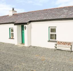 Exterior of a white cottage with green door and windows with two benches in a gravel yard at Pat White's Cottage in Rostrevor