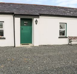 A white cottage exterior with a green door two windows a bench and gravel ground at Pat Whites Cottage in Rostrevor