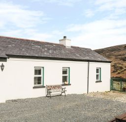 A white cottage with a green door and three windows with a bench outside on gravel at Pat White's Cottage in Rostrevor