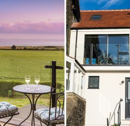 A balcony with a small round table and two chairs overlooking a green field and the sea at Emblestones in Embleton